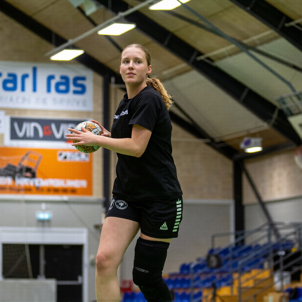 Kvindelig håndboldspiller i sort holder håndbold i hal